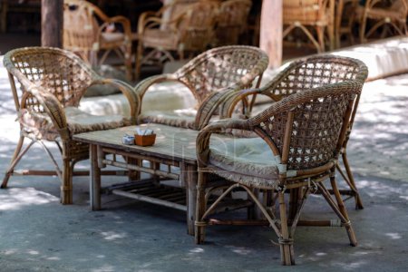 Cadre extérieur avec trois chaises en osier et une table assortie ornée de coussins. Le mobilier est disposé sur un sol en béton sous l'ombre, créant une atmosphère accueillante pour la détente
