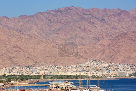 Photo for A coastal city nestled at the foot of a massive rugged mountain range in Elam, Israel. The citys architecture is dense with buildings closely packed together. Boats are docked near the shore a port - Royalty Free Image