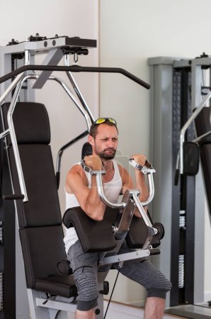 A man exercising in a gym. Male using a machine for upper body workouts and they are wearing sunglasses a white tank top, and grey shorts. The gym appears well-equipped. Strength and motivation, sport