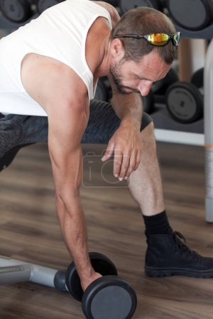 An individual is engrossed in a workout, lifting dumbbells. They sport sunglasses, a white top, and dark pants. The gym is well equipped. Close up handsome middle aged man doing dumbbell. Working out