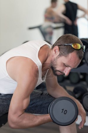 Skinny guy hold, doing a dumbbells up in hand. Thin man in sports with dumbbells. Weak man lift a weight, biceps, muscle, fitness. Man holding dumbbell in hand sunglasses, a white top and dark short