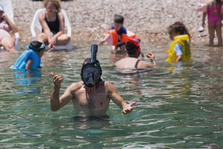 Téléchargez les photos : On voit un homme torse nu portant un masque et un tube de plongée, nageant tranquillement dans les eaux claires de la mer. Entouré d'enfants et d'adultes pataugeant et jouant dans les eaux peu profondes, la chaude journée d'été - en image libre de droit