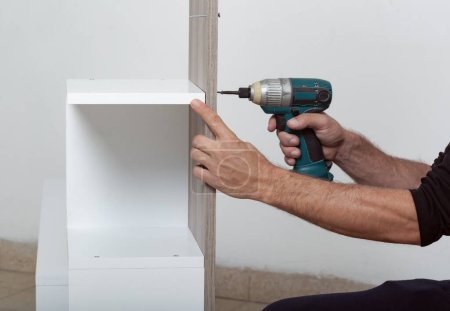 Téléchargez les photos : Un homme assemblant une étagère blanche. Une perceuse électrique est utilisée pour fixer une planche de bois à l'étagère. Les mains de mans sont visibles, indiquant le processus d'assemblage. L'étagère est contre un mur blanc. - en image libre de droit