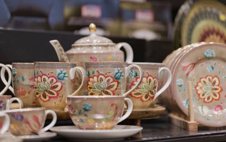A porcelain tea set with intricate floral designs against a dark background. The set includes cups, saucers, a teapot, and plates. Each piece is adorned with vibrant artwork of flowers and leaves.