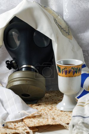 Máscara de gas, parcialmente oculta por la oración blanca talit, junto a una taza de Kadish en una mesa con pan matzá. Fiesta del vino matzoth celebración matzoh pan de Pascua judía. Elementos tradicionales y supervivencia