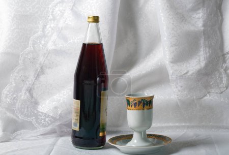 Photo for Bottle is dark with a golden cap of grape juice and a Kiddush cup against a white lacy curtain. The cup has colorful designs and sits on a matching saucer. Shabbat, passover, pesah celebration concept - Royalty Free Image