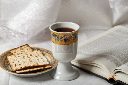 Coupe à vin casher et pain matsa placés à côté d'un livre ouvert. La toile de fond est un élégant tissu blanc. Coupe est ornée de dessins complexes et le matzah repose sur une plaque décorative. Pessah juif