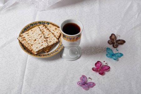 Matsa, pain sans levain sur une assiette et coupe de vin décorées de façon ornementale. Quatre papillons ajoutent une touche de fantaisie. Le décor de la Pâque est sur une nappe blanche à motifs subtils. Judaïsme, Religieux