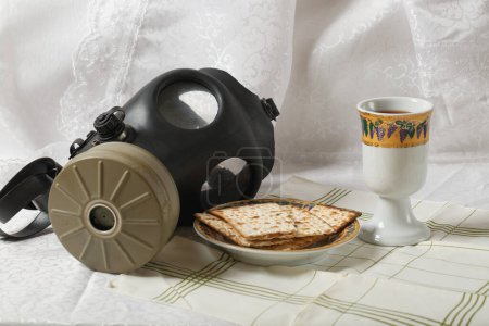 Foto de Máscara de gas, matzá en un plato y una taza adornada en una mesa con un paño a rayas. El telón de fondo es una cortina de encaje blanco. La máscara de gas contrasta con la comida y la bebida. Mascara de gas israelí. Ataque radiactivo - Imagen libre de derechos