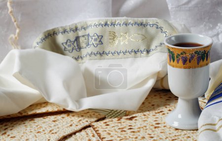 Foto de Talit blanco con bordado azul doblado junto a trozos de matzá y copa Kidush llena de vino. Las piezas de Matzah son planas y tienen un patrón perforado. La copa Kidush, tallit. Celebración religiosa - Imagen libre de derechos