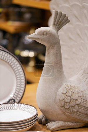 Figurine de paon en céramique en blanc, exposée bien en vue parmi les assiettes. Le paon, avec des plumes détaillées et une posture élégante, se distingue. Le fond révèle des étagères avec divers articles de cuisine