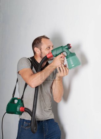 Un hombre posando humorísticamente con un pulverizador de pintura como si fuera una pistola. Está vestido con atuendo casual, vaqueros azules y una camiseta corta gris contra una pared blanca. El pulverizador, de color verde, manguera negra
