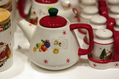 Variedad de teteras y recipientes de cerámica, todos adornados con diseños festivos. Una tetera con una base blanca y coloridos motivos navideños está en primer plano, incluyen Santa Claus, regalos, campanas y copos de nieve.