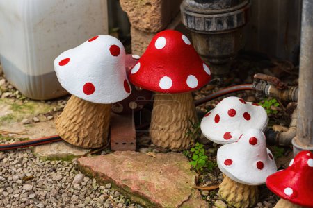 Several brightly painted figurine of a fly agaric, featuring red and white polka dots, are set amidst garden pathways, surrounding area includes rocks, gravel and scattered greenery. Garden decoration