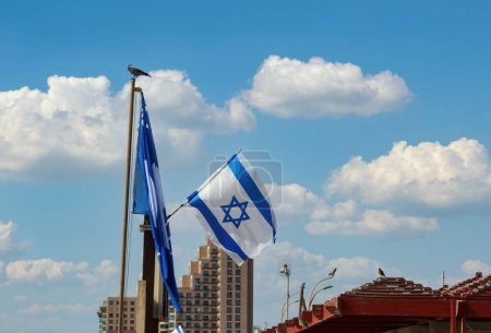 Two Israeli flags flutter atop poles against a clear blue sky with scattered white clouds. Buildings and a bird are visible in the background. Israel flags with a star of David over sky background.