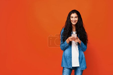 Foto de Mujer árabe atractiva joven de pie con teléfono inteligente en las manos. Aislado en estudio sobre fondo naranja. - Imagen libre de derechos