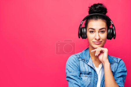 Foto de Retrato de una joven mujer árabe atractiva de pie en el estudio con auriculares. Disfrutando escuchando música. Chica en ropa casual aislada sobre fondo rosa - Imagen libre de derechos
