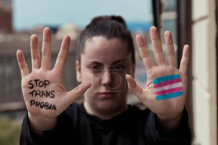 Foto de Una chica con las manos frente a ella. Ella tiene una mano pintada con la bandera trans y por el otro el mensaje stop transpohia - Imagen libre de derechos