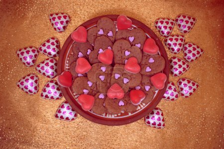 Vegetarian cookies of different types of hearts made using plungers. Decorated with natural juices thickened with agar-agar. Chocolate candies in the shape of hearts in wrappers of red and mixed colors. Gold background.