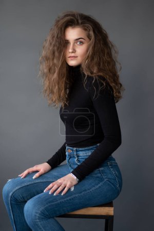 Foto de Sensual girl with curly hair, sitting on high chair, on a grey background, looking at camera. - Imagen libre de derechos