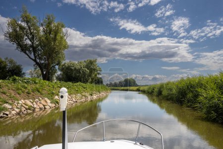 Téléchargez les photos : Canal built between Morava part of Czech and Slovakia by Bata. Beautiful greenery lining the route, trees, bushes, grass and flowers. Blue sky with white clouds. Part of the boat visible. Bata canal, Czech and Slovak republics - en image libre de droit