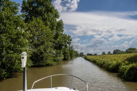 Téléchargez les photos : Canal built between Morava part of Czech and Slovakia by Bata. Beautiful greenery lining the route, trees, bushes, grass and flowers. Blue sky with white clouds. Part of the boat visible. Bata canal, Czech and Slovak republics - en image libre de droit