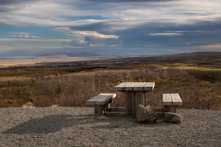 Foto de Country with trees, bushes, grass. Mountains on the horizon. Natural wooden desk and benches to sit, relax, eat and enjoy the panorama. Intense clouds on the sky. Reykhold, south-middle Iceland - Imagen libre de derechos