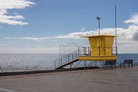 Photo for Promenade and lifeguard yellow tower on the beach beside. Calm water of Atlantic ocean in the winter. Blue sky with white clouds. Tarajalejo, Fuerteventura, Canary Islands, Spain. - Royalty Free Image