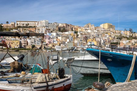 Long pier with various sizes and colors of fishing boats. In the background houses of the city built up the hill. Sciacca, Sicily, Italy.