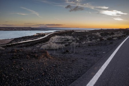 Foto de Empty good quality asphalt road on the coastline, crossing lava field. Sun on the horizon during sunset. Blue sky with colorful clouds. Salinas de Janubio, Lanzarote, Canary Islands, Spain. - Imagen libre de derechos