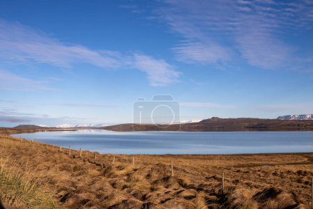 Trockenes Gras auf den Feldern und Weiden. Ruhiges Wasser der Fjorde. Berge mit Schnee am Horizont. Blauer Himmel mit weißen Wolken. Sonniger Tag. Gebiet von Hvitserkur, Nordwest-Island.