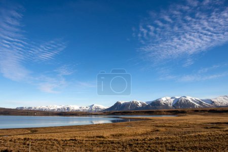 Hierba seca en los campos y pastos. Aguas tranquilas de los fjors. Montañas con nieve en el horizonte. Cielo azul con nubes blancas. Día soleado. Zona de Hvitserkur, noroeste de Islandia.