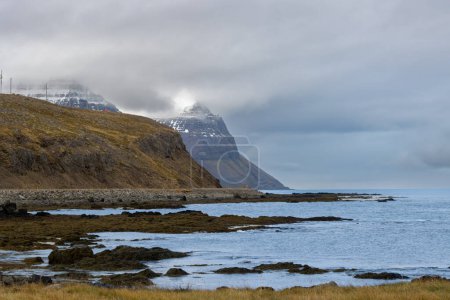 Wilde Küste des Fjordgebiets Westfjords. Ruhiges Wasser des Atlantiks und majestätische Berge mit etwas Schnee im Herbst. Bewölkter Himmel. Gebiet von Isafjordur, Nordwest-Island.