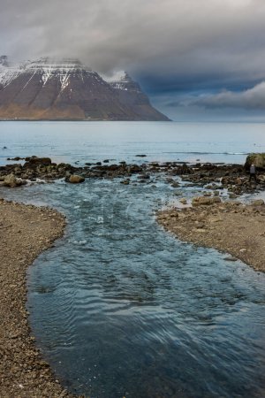 Wilde Küste des Fjordgebiets Westfjords. Ruhiges Wasser des Atlantiks und majestätische Berge mit etwas Schnee im Herbst. Bewölkter Himmel. Gebiet von Isafjordur, Nordwest-Island.