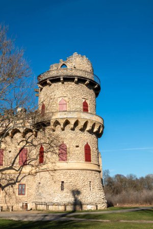 Pseudo-gothic imitation of a castle ruin, made of stone. Located in a park with green grass in the late winter and many trees. Bright blue sky. Red window shutters. UNESCO heritage. Januv hrad, Podivin, Lednice, Morava, Czech republic.