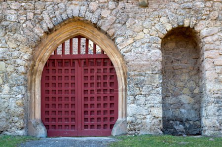 Stone wall of a castle. Wooden arch of a gate. Square structure of a bright red gate. Bright green grass in the late winter. Januv Hrad, Lednice, Podivin, Moravia, Czech republic.