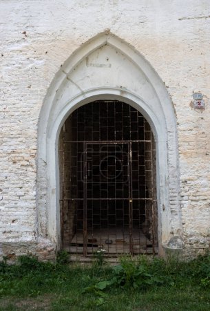 Pared de baldosas, pintada de blanco con una entrada de arco. Red oxidada de metal. Hierba verde brillante. Ruina de una mansión histórica, Breclav, Moravia, República Checa.
