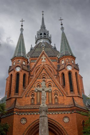 Edificio de una iglesia neogótica de Visitación de la Virgen María, hecha de ladrillos naranjas. Estatua de la Crucifixión en el parque. Postorna, Breclav, Moravia, República Checa.