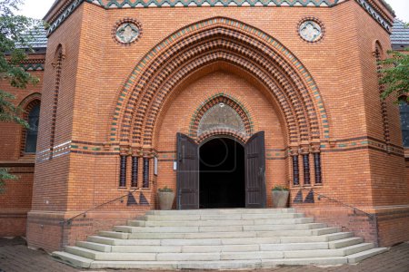 Haupteingang zum Gebäude der neugotischen Mariä Heimsuchung-Kirche aus orangefarbenen Ziegeln. Weit geöffnetes dunkelbraunes Holztor. Postorna, Breclav, Mähren, Tschechische Republik.