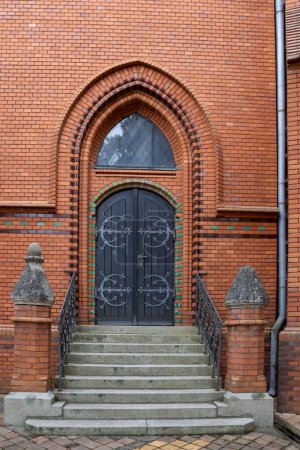 Side door to the building of a neo-gothic church of Visitation of Virgin Mary, made of orange bricks. Wide opened dark brown wooden gate. Postorna, Breclav, Moravia, Czech republic.