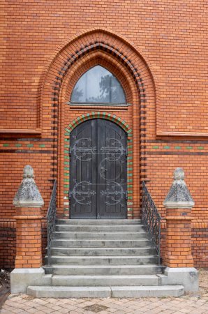 Puerta lateral al edificio de una iglesia neogótica de Visitación de la Virgen María, hecha de ladrillos naranjas. Puerta de madera marrón oscuro abierta. Postorna, Breclav, Moravia, República Checa.