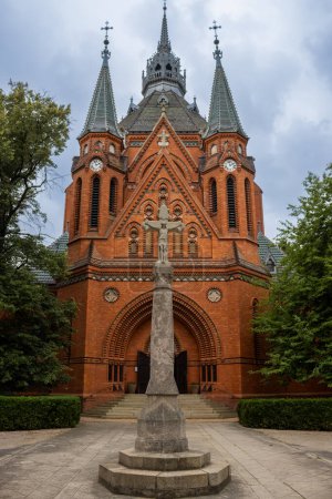Edificio de una iglesia neogótica de Visitación de la Virgen María, hecha de ladrillos naranjas. Estatua de la Crucifixión en el parque. Postorna, Breclav, Moravia, República Checa.