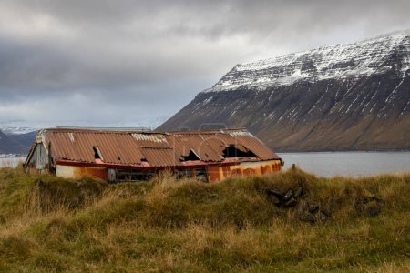 Verlassenes Haus mit einem rostigen und kaputten Dach. Herbstgras liegt herum. Das Hotel liegt an der Küste eines Fjords mit einem großen Berg an der anderen Küste, mit Schnee auf der Spitze. Gebiet von Isafjordur, Westfjorde, Island.