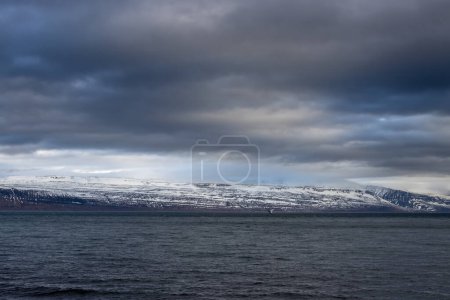 Calm water of Atlantic ocean. High mountains on the coast in the background. Intense cloudy sky in the autumn. Area of Isafjordur, Westfjords, Iceland.