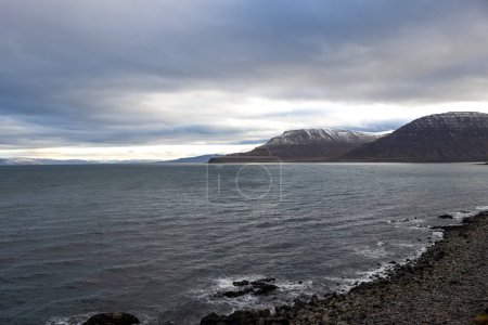 Calm water of Atlantic ocean. High mountains on the coast on the other side of fjord. Rocky coast in the foreground. Intense cloudy sky in the autumn. Area of Isafjordur, Westfjords, Iceland.