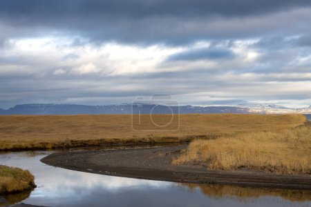 Ruhiges Wasser des Flusses, das den bewölkten Himmel mit einem Hauch von Sonnenlicht reflektiert. Gelbes trockenes Herbstgras liegt herum. Punkt, bevor der Fluss in den Ozean fließt. Westfjord, Island.