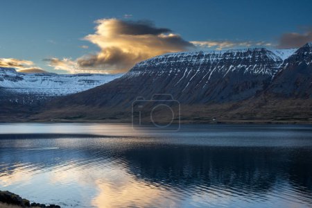 Die schneebedeckten Berge. Bunte Wolken am blauen Himmel bei Sonnenuntergang Ruhiges Wasser des Atlantiks im Fjord. Gebiet der Westfjorde, Isafjordur, Island.
