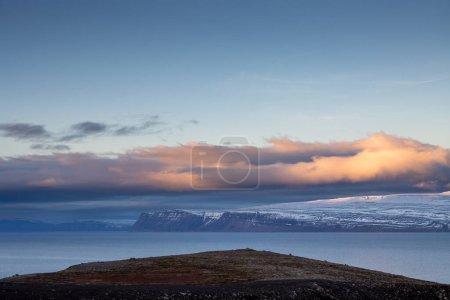 Die schneebedeckten Berge. Bunte Wolken am blauen Himmel bei Sonnenuntergang Ruhiges Wasser des Atlantiks im Fjord. Gebiet der Westfjorde, Isafjordur, Island.