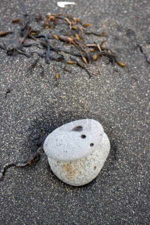 Two bigger pebbles one on the other. Three holes - dots on the top one. Black volcanic sand on the beach. Dry algae in the blurry background. Area of Isafjordur, Westfjords, Iceland.