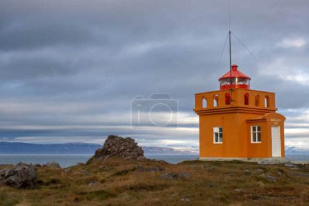 Leuchtend orange Fassade des Leuchtturms. Roter Turm mit dem Licht. Bewölkter Himmel im Herbst. Gebaut an der Küste des Atlantiks in der Gegend von Isafjordur, Westfjorde, Island.